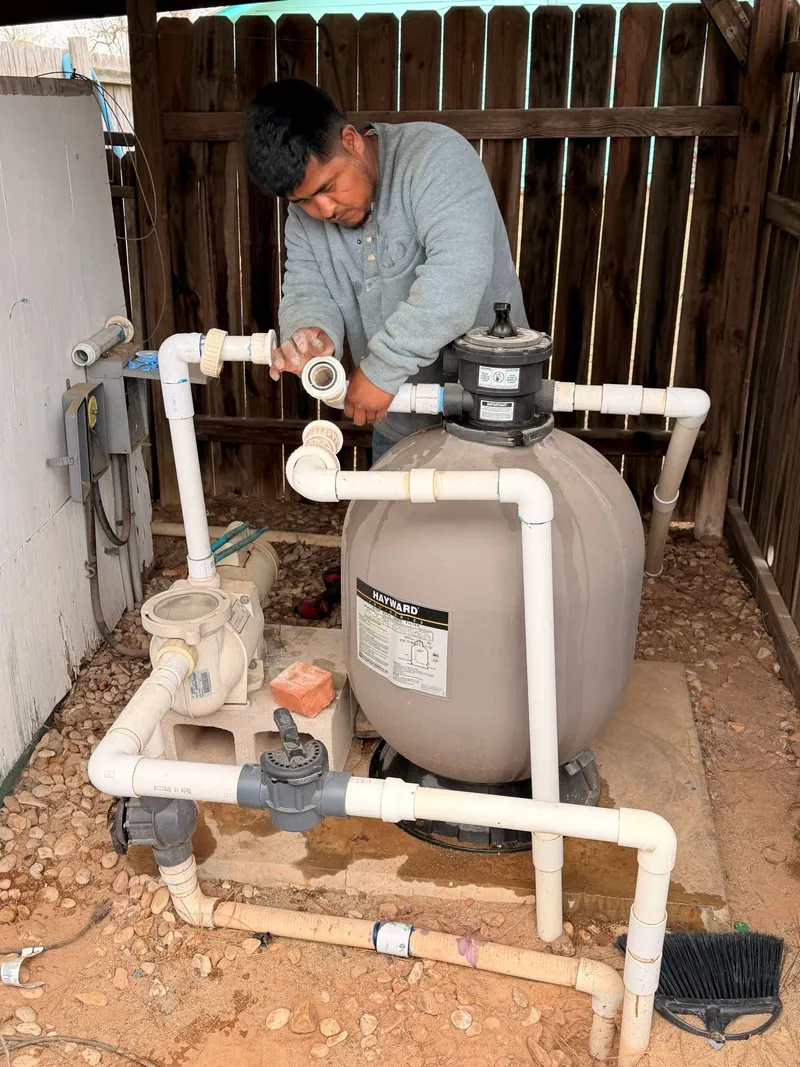 Crew member replumbing white PVC pipe between a pool pump and a sand filter on the equipment pad